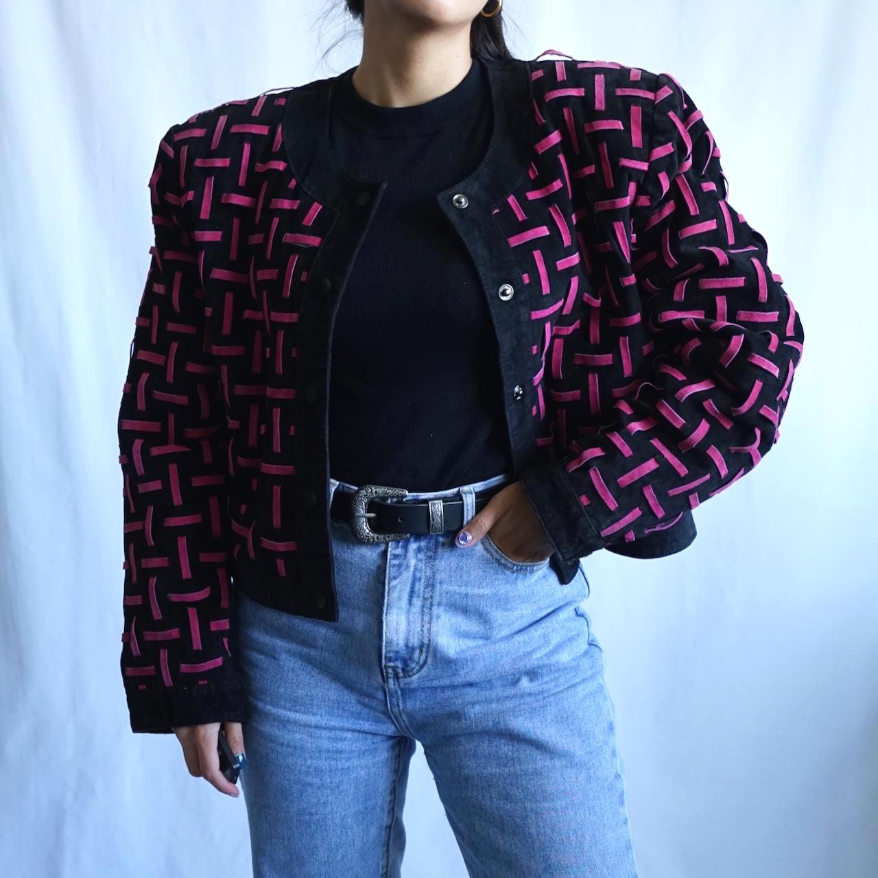 A person wearing a black cropped jacket with a pink geometric woven pattern, styled over a black top and light blue jeans against a light blue backdrop.