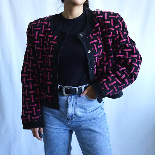 A person wearing a black cropped jacket with a pink geometric woven pattern, styled over a black top and light blue jeans against a light blue backdrop.